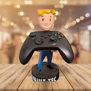 May include: A black video game controller held by a figurine with a smiling face and blonde hair. The figurine is wearing blue pants and standing on a black base with "VAULT TEC" printed on it. The controller has colored buttons.