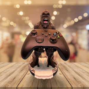 May include: A brown gorilla figurine holding a black Xbox controller. The gorilla has an open mouth and is set on a base with a water-like design. The controller has colored buttons.