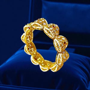 Pode incluir: Uma pulseira de ouro com intrincados detalhes de filigrana e detalhes de pedras preciosas. A pulseira apresenta uma série de secções ornamentadas e conectadas, cada uma adornada com pedras preciosas claras e vermelhas. A pulseira é apresentada numa superfície de veludo azul.