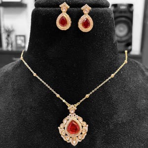 Peut inclure: Un ensemble de bijoux doré comprenant un collier et des boucles d'oreilles assorties. Le collier présente un pendentif en forme de goutte d'eau, avec une pierre rouge entourée de petites pierres claires. Les boucles d'oreilles reprennent le design du pendentif. L'ensemble est présenté sur un fond noir.