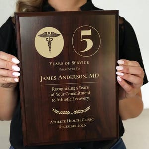 May include: A dark wood plaque with gold lettering and accents, held by a person. The plaque reads "5 Years of Service" and is presented to "James Anderson, MD." It recognizes commitment to athletic recovery, with the Athlete Health Clinic, December 2026.