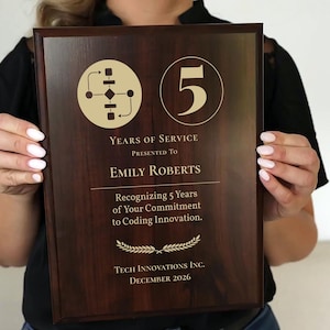 May include: A dark wood plaque with gold lettering and graphics. The plaque reads "5 Years of Service" and is presented to "Emily Roberts." It recognizes her commitment to coding innovation. The bottom of the plaque includes the company name and the date, December 2026.