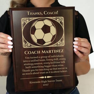 May include: A wooden plaque with a soccer ball design and the text "Thanks, Coach!" is held by a person. The plaque includes the name "COACH MARTINEZ" and a message from the Riverside High Soccer Team, dated 2027.