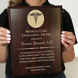 May include: A dark brown wooden plaque with gold lettering. The plaque reads "Medical Care Excellence Award" presented to "Thomas Bennett, MD". The plaque also includes a gold medical symbol and the text "In recognition of your excellence in the medical field. Your expertise and caring approach have made a lasting impact on the lives of many. Greenwood Medical Center December 2026."