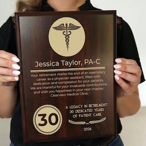 May include: A dark brown wooden plaque with gold lettering and a gold medical symbol. The plaque reads "Jessica Taylor, PA-C" and "A Legacy in Retirement: 30 Dedicated Years of Patient Care".