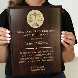 May include: A wooden plaque with the text "ACCURATE TRANSCRIPTION EXCELLENCE AWARD" and the name "Laura Mitchell". The plaque features a gold scale icon and is presented by Clark County in December 2026. The plaque is held by a person.