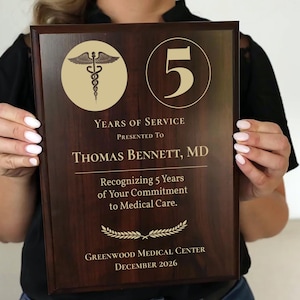 May include: A dark brown wooden plaque with gold lettering and a gold medical symbol. The plaque reads "5 Years of Service Presented to Thomas Bennett, MD Recognizing 5 Years of Your Commitment to Medical Care. Greenwood Medical Center December 2026"