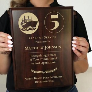 May include: A rectangular dark wood plaque featuring a gold-colored graphic of a ship, the number 5, and text reading: Years of Service, Presented To Matthew Johnson, Recognizing 5 Years of Your Commitment to Port Operations, North Beach Port Authority, December 2026.