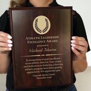 May include: A wooden plaque with the text "ATHLETIC LEADERSHIP EXCELLENCE AWARD" presented to Michael Johnson. The plaque has a gold emblem and includes text recognizing excellence in athletic direction. The Citywide Sports Center presented the award in December 2026.
