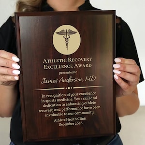 May include: A dark wood plaque with gold lettering and a medical symbol. The plaque reads "ATHLETIC RECOVERY EXCELLENCE AWARD" and is presented to James Anderson, MD. The text also mentions the Athlete Health Clinic and December 2026.