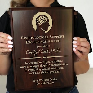 May include: A wooden plaque with the text "PSYCHOLOGICAL SUPPORT EXCELLENCE AWARD" and the name "Emily Clark, Ph.D." is held by a person. The plaque has a gold-colored brain and profile graphic. The text on the plaque also mentions "Total Wellness Center" and "December 2026."
