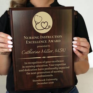 May include: A dark brown wooden plaque with gold lettering. The plaque has a stethoscope and heart design in the top left corner. The text reads "Nursing Instruction Excellence Award presented to Catherine Miller, MSN. In recognition of your excellence in nursing education. Your expertise and dedication have profoundly impacted the next generation of nursing professionals. Stonebrook University December 2026."