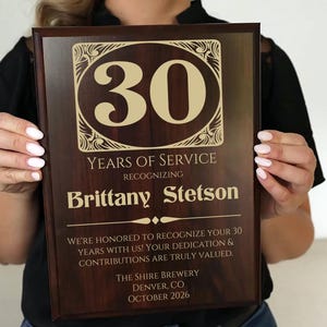 May include: A brown wooden plaque with gold lettering recognizing 30 years of service for Brittany Stetson. The plaque features a decorative border and the text "We're honored to recognize your 30 years with us! Your dedication & contributions are truly valued. The Shire Brewery, Denver CO, October 2026."