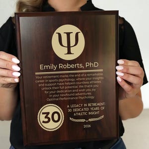 May include: A wooden plaque with a dark brown finish, featuring gold text and a circular emblem. The plaque commemorates Emily Roberts, PhD, for 30 years of athletic insight. The text includes the year 2026.