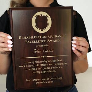 May include: A rectangular wooden plaque with the text "REHABILITATION GUIDANCE EXCELLENCE AWARD" and "presented to John Davis". The plaque includes a shield emblem and text recognizing the recipient's work as a parole officer. The plaque is dated December 2026.