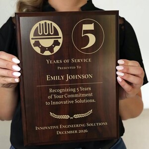 May include: A wooden plaque with gold lettering and a dark brown finish. The plaque reads "Years of Service" and "Emily Johnson." It also includes the text "Recognizing 5 Years of Your Commitment to Innovative Solutions." The date "December 2026" is also visible.
