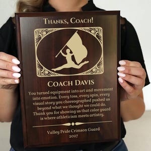 May include: A wooden plaque with the text "Thanks, Coach!" and "Coach Davis" in gold lettering. The plaque features a gold illustration of a person with a flag. The text below reads "Valley Pride Crimson Guard 2027".