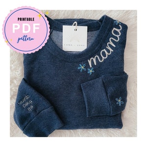 Op de afbeelding: Een donkerblauw sweatshirt met het woord "mama" geborduurd in wit op de voorkant. Het woord is omringd door kleine blauwe bloemen. Het sweatshirt heeft ook de woorden "Luna June" op een wit label dat aan de halslijn is bevestigd.