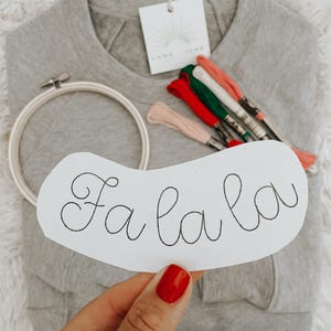 May include: A white embroidery hoop with a piece of paper inside. The paper has the words "Fa la la" written in cursive. There are also several spools of embroidery floss in red, green, and pink.