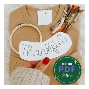 May include: A craft kit with a tan-colored sweatshirt, embroidery floss, and a wooden hoop. A white card with the word "Thankful" is held in front of the sweatshirt. A green and purple "Printable PDF Pattern" sticker is in the lower right corner.