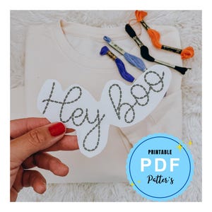 May include: Printable PDF pattern for embroidery, featuring the text "Hey boo" in a cursive font. The pattern is on a white background and is being held by a hand.