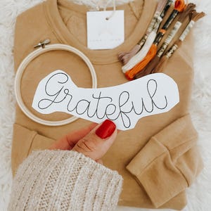 May include: A tan sweatshirt with a white tag that says "Love & Luxe" and a white paper with the word "Grateful" written in black cursive. There is a wooden embroidery hoop and a bundle of embroidery floss in various colors.