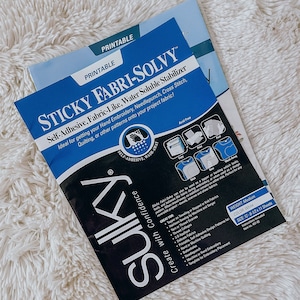 May include: A blue package of Sulky Sticky Fabri-Solvy water soluble stabilizer for embroidery, quilting, and needlepunch. The package is printed with instructions and features a hand washing away icon. The text on the package reads "Self-Adhesive, Fabric-Like, Water Soluble Stabilizer" and "Create with Confidence".