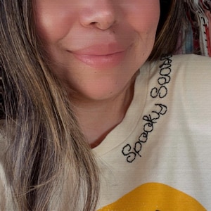 May include: Cream-colored t-shirt with a black cat graphic and a yellow moon. The words "Spooky Season" are embroidered in black on the neckline. The person in the image is smiling.