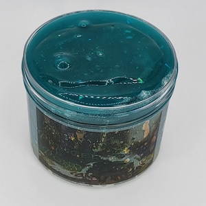 May include: A clear plastic container filled with teal blue slime with iridescent glitter. The slime is thick and gooey and has a smooth texture.