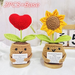 May include: Two crochet "Emotional Support Plants" are displayed. One features a red heart, the other a yellow sunflower. Both have green leaves and are in brown pots with smiling faces. Each holds a card with text.