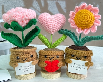 May include: Three crocheted potted plants with cute faces. The plants are a pink flower, a pink heart, and a yellow flower with pink petals. Each pot has a message that says "Thank you for helping me grow".