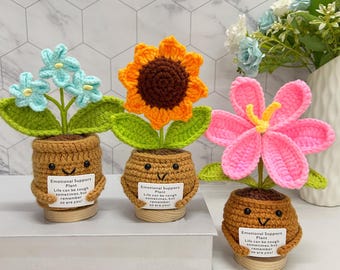 May include: Three handmade crochet "Emotional Support Plants" in brown pots with smiling faces. One has blue flowers, one a sunflower, and the last a pink flower. Each pot has a tag with text: "Life can be tough sometimes, but remember so are you!"