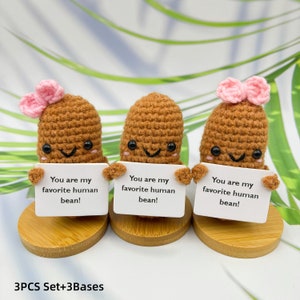 May include: Set of three crocheted brown bean-shaped figures with pink bows, each holding a white sign that reads "You are my favorite human bean!" The figures are standing on small wooden bases.