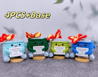 Peut inclure: Quatre peluches en crochet en forme de bennes à ordures avec des flammes sur le dessus. Chaque benne à ordures tient une pancarte qui dit "Emotional Support Dumpster Fire. It's fine. I'm fine. Everything is fine!" Les jouets sont posés sur des bases en bois.