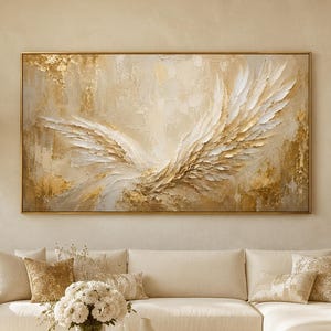 May include: A large rectangular wall art piece featuring an abstract painting of white and gold wings with textured brushstrokes. The artwork is framed in a thin gold border and displayed above a cream-coloured sectional sofa in a bright room.