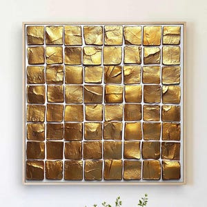 Könnte beinhalten: Ein quadratisches, gerahmtes Kunstwerk mit einem Raster aus strukturierten, goldfarbenen Quadraten. Das Kunstwerk hängt an einer weißen Wand über einem hellen Holztisch. Auf dem Tisch stehen eine blaue Ablage, Bücher und eine Teekanne.