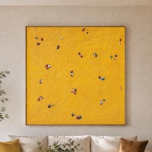 Puede incluir: Un gran cuadro cuadrado sobre lienzo con un fondo amarillo vibrante y texturizado, adornado con pequeñas figuras abstractas multicolores dispersas. La obra se exhibe en una pared clara sobre un sofá de tonos neutros y una mesa de centro redonda de madera.
