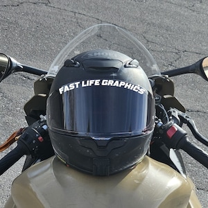 Puede incluir: Un casco de moto negro con una visera transparente y una pegatina blanca que dice "FAST LIFE GRAPHICS". El casco está sentado en una motocicleta.