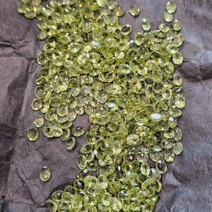 Puede incluir: Una colección de gemas ovaladas de color verde claro dispersas sobre una superficie oscura y texturizada. Las gemas están facetadas, reflejando la luz y creando un efecto brillante. La impresión general es de abundancia y lujo.