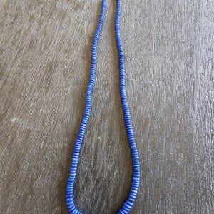 Puede incluir: Un collar largo de cuentas azules. El collar está compuesto por numerosas cuentas pequeñas y circulares ensartadas, creando un bucle continuo. Las cuentas son de tamaño y color uniformes, dando al collar un aspecto elegante y moderno.