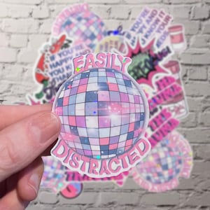 Peut inclure: Un autocollant holographique avec un motif de boule disco et les mots "EASILY DISTRACTED" en lettres roses à bulles. La boule disco est composée de carrés roses, bleus et violets. D'autres autocollants avec du texte et des graphiques sont en arrière-plan.