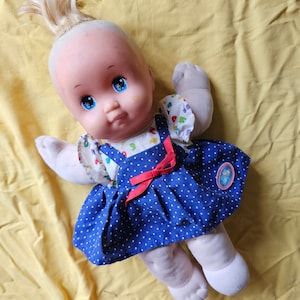 May include: A vintage baby doll with blonde hair and blue eyes. The doll is dressed in a blue polka-dot dress with a red bow and a white floral print top. It rests on a yellow surface.