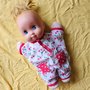 May include: A doll with blonde hair wearing white and red cherry print pyjamas. The doll has blue eyes and a rosy complexion.