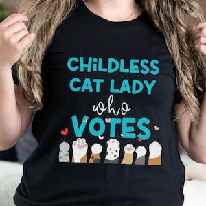 May include: Black t-shirt with the text "Childless Cat Lady who Votes" in teal blue lettering. The shirt features a row of colorful paw prints with hearts.