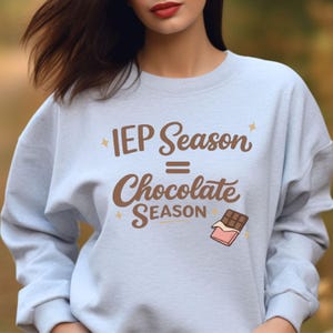 Könnte beinhalten: Hellblaues Sweatshirt mit dem Text "IEP Season = Chocolate Season" in brauner Schrift. Eine Grafik einer Schokoladentafel mit rosa Verpackung befindet sich darunter. Das Sweatshirt hat einen Rundhalsausschnitt und lange Ärmel.