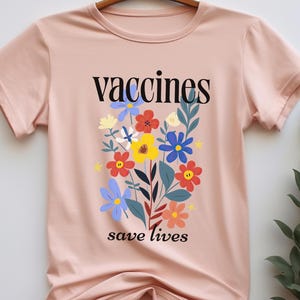 May include: A light pink t-shirt with the words "vaccines save lives" in black text. The shirt features a colorful floral design with red, yellow, blue, and white flowers.