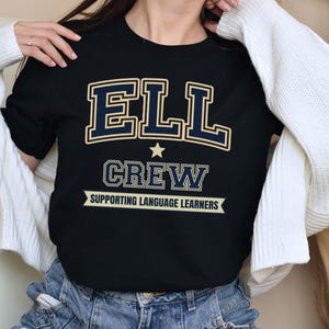 May include: Black crew neck t-shirt with the text "ELL CREW SUPPORTING LANGUAGE LEARNERS" in navy blue and gold lettering. A star is above the word "CREW". The shirt is worn with blue jeans and a white cardigan.
