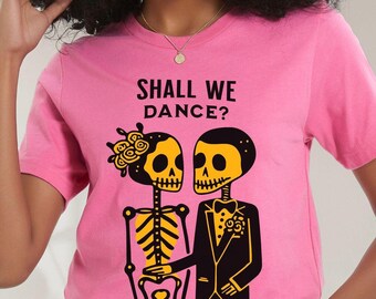 May include: A bright pink short-sleeve t-shirt featuring a graphic of two skeletons in yellow and black. The skeleton on the left wears flowers, and the one on the right wears a tuxedo. The text "SHALL WE DANCE?" is printed in black above them.