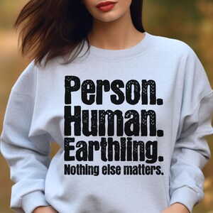 Op de afbeelding: Een lichtblauw sweatshirt met ronde hals en zwarte tekst: Person. Human. Earthling. Nothing else matters. De letters hebben een verweerde, getextureerde uitstraling en het kledingstuk heeft lange mouwen en een geribbelde halslijn.