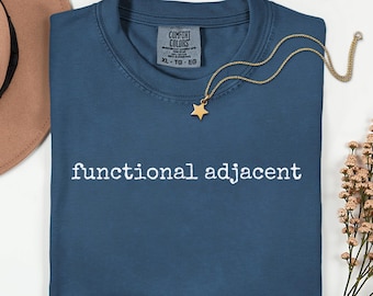 May include: A blue t-shirt with the words "functional adjacent" in white text. A gold star pendant necklace rests on the shirt. The shirt has a Comfort Colors tag, and a brown hat is partially visible.